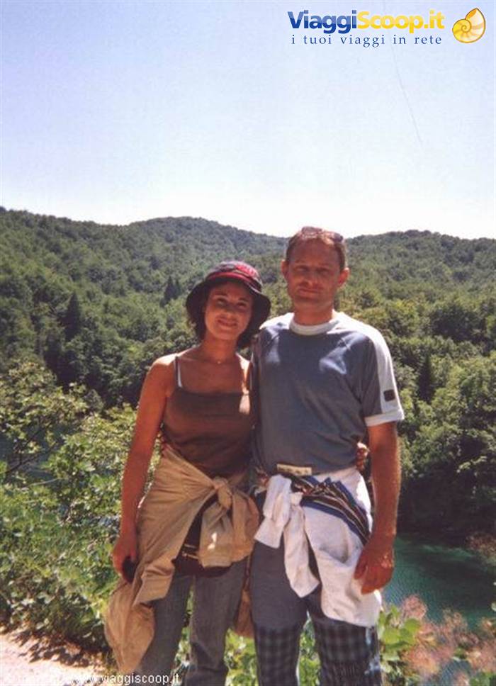 smiling couple in hat and t-shirt beside a turquoise lake surrounded by green woods