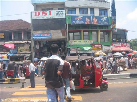 Pedestrians cross the street near shops, a red tuk-tuk drives through the crowd