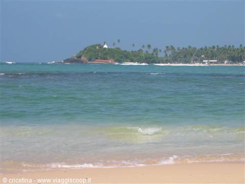 Turquoise sea in front of a palm-covered islet with white building, sandy beach