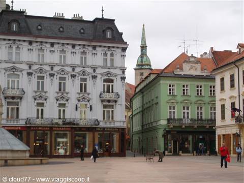 Square with colorful historic buildings and green bell tower, pedestrians under grey sky