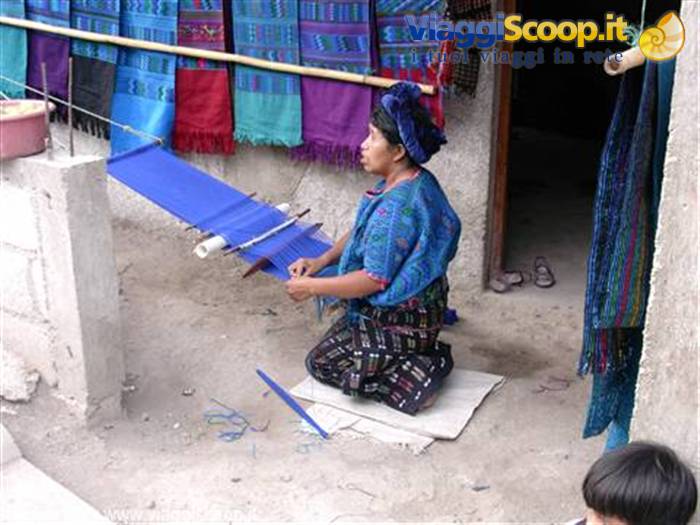 Maya woman weaves blue fabric on backstrap loom, colorful textiles hanging behind