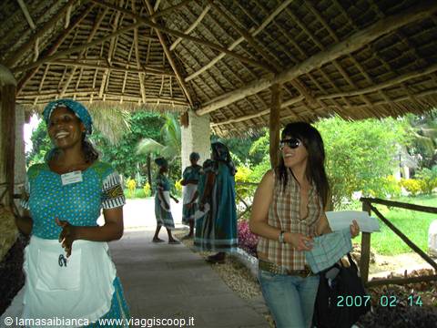 Smiling woman in white apron chats with a tourist under a thatched roof