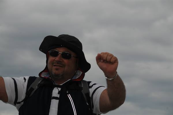 Man in black hat, sunglasses and backpack raises his fist under cloudy sky