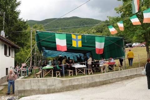 Outdoor festival under green tent with Italian flags, wooden tables, wooded hills