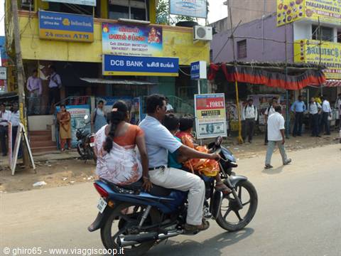 Family of three riding a motorbike past shops on an Indian street