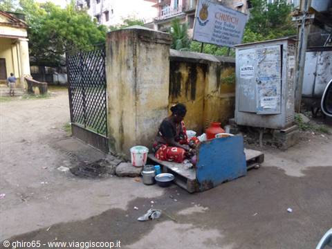 Woman sitting with stoves and pots beside a weathered yellow wall