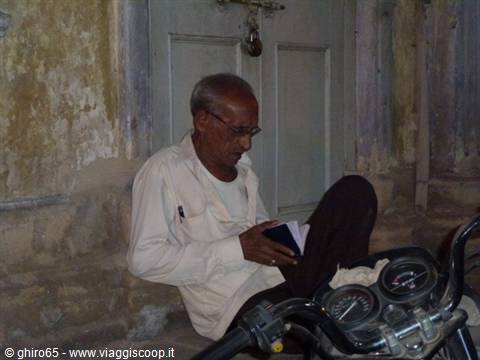 Elderly man reads a notebook beside a motorcycle in front of a peeling door