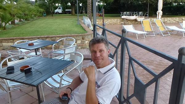 Smiling man seated on terrace with metal tables and loungers near the pool