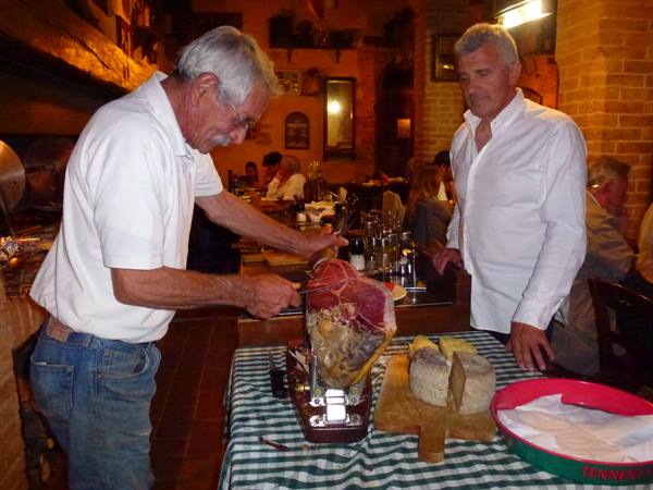 Two men slicing prosciutto with cheese on the table in a trattoria