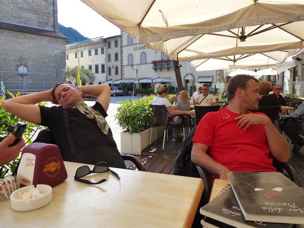 Relaxed tourists at Bar della Piazza tables under umbrellas, square with old buildings