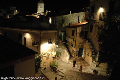 Night alley in old stone village, lit bell tower, people on the steps