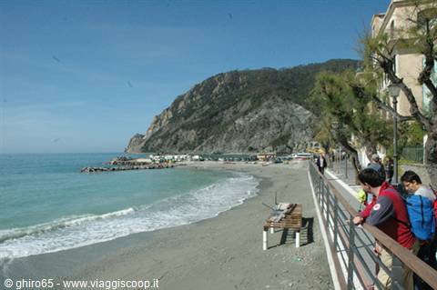 Tourists on a railing walkway overlooking Monterosso sandy beach, mountain behind