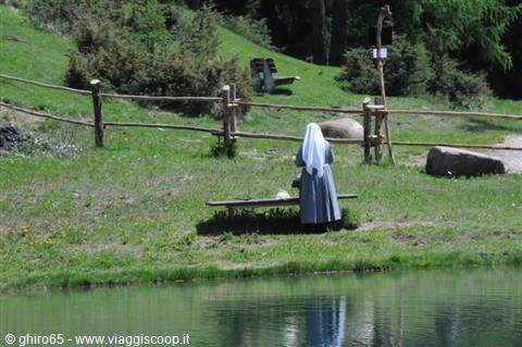 A nun in a blue habit sitting on a wooden bench by an alpine pond