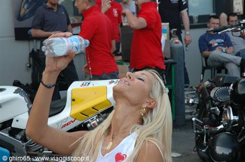 Blonde woman drinking water beside race bikes and mechanics in red shirts