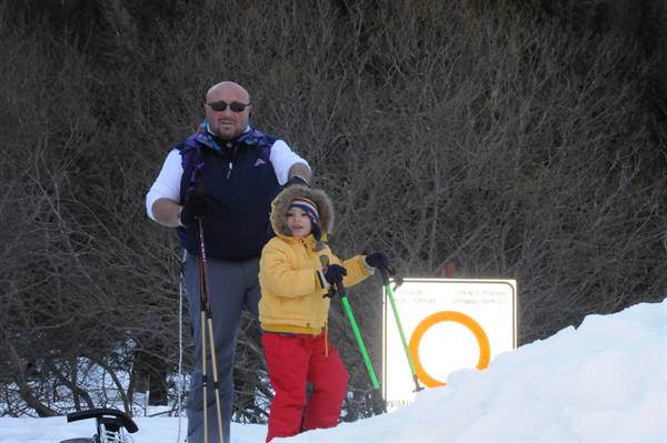 Father and child holding ski poles beside a trail sign in the snow