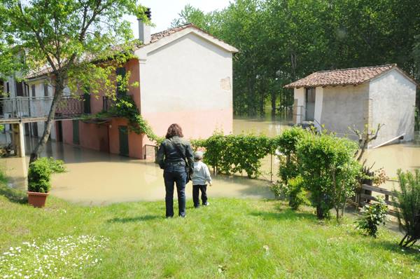 Woman and child watch brown floodwater surrounding rural houses and garden