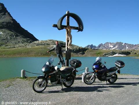 Man beside a wooden and stone crucifix between two motorcycles by a green alpine lake