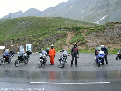 Motorcyclists in helmets beside parked bikes on wet asphalt among grassy mountains