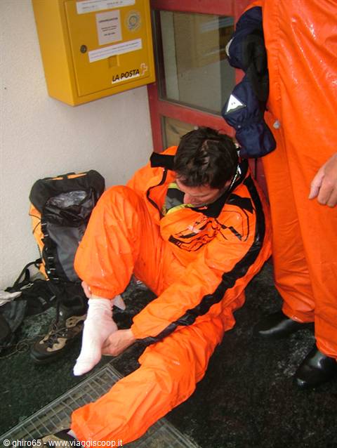 Man in wet orange rain suit removing his boot beside a yellow La Posta box