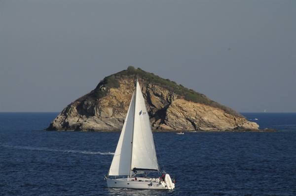 White sailboat sailing in front of a rocky islet covered with greenery