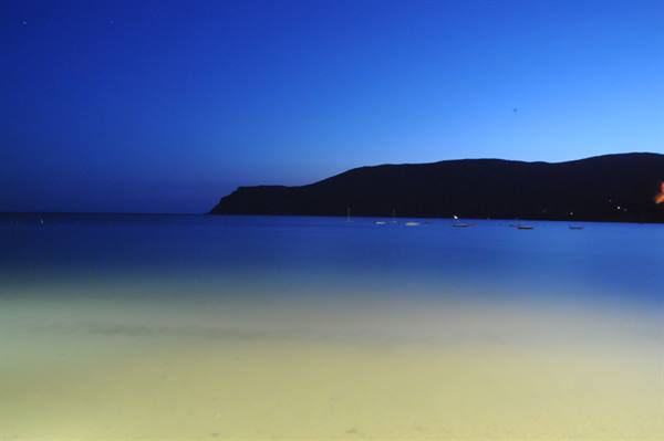 Night sandy beach, calm sea and dark hill under blue sky
