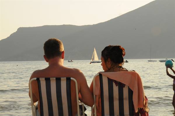 Couple on striped deckchairs watching a sailboat on calm sea