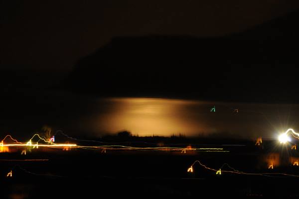 Car light trails along night shore, golden glow reflecting on dark water
