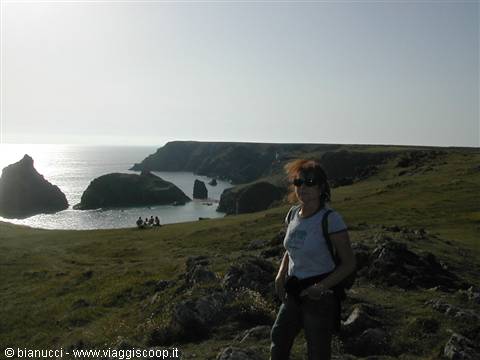Woman with backpack on grassy cliffs, sea stacks and beach at Kynance Cove