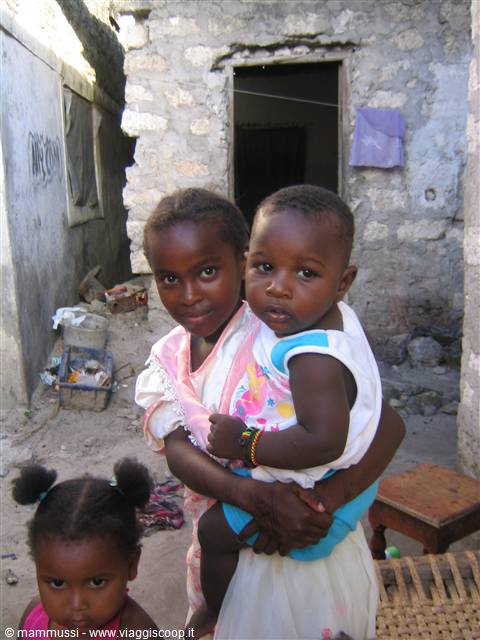 Young girl holding a baby in front of stone house, another child watching