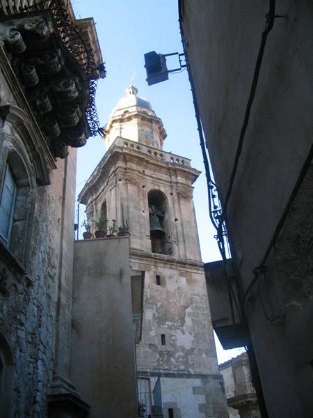 Stone bell tower with bells between old buildings, blue sky