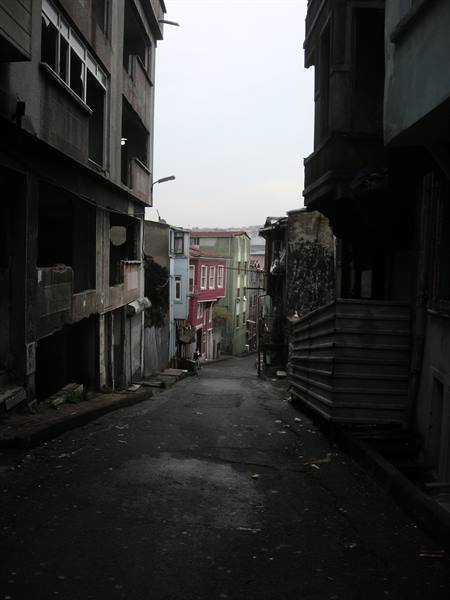 narrow descending street between decaying buildings, a streetlamp and grey sky