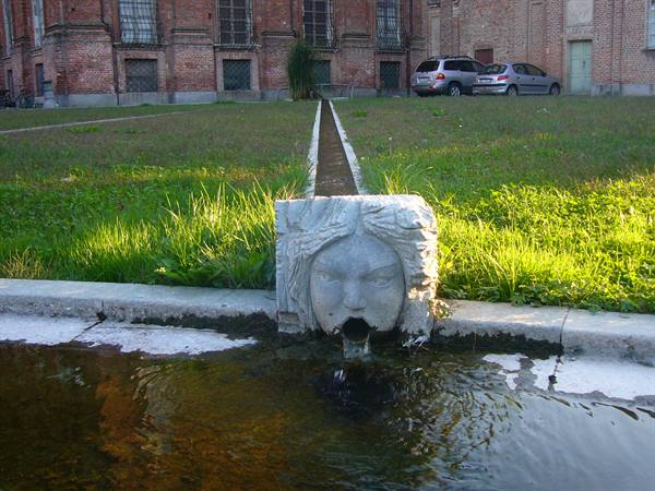 Stone fountain with carved face pouring water, lawn and brick building behind
