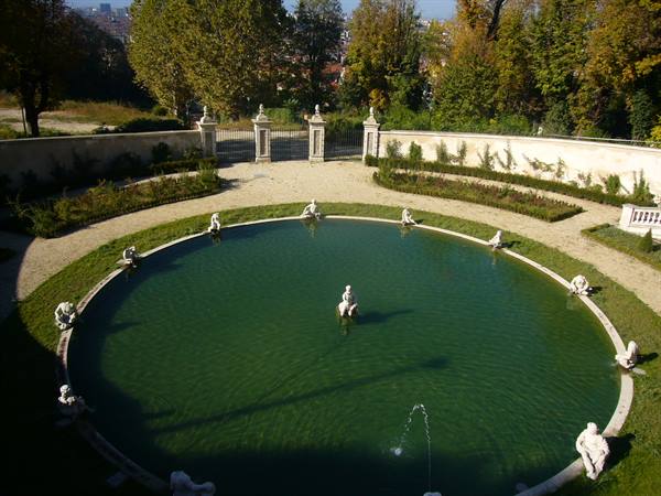 Circular green pond with white statues and water jet, baroque garden in sunlight