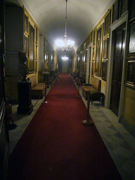 Gallery hallway with red carpet, gilt paintings, busts and chandeliers