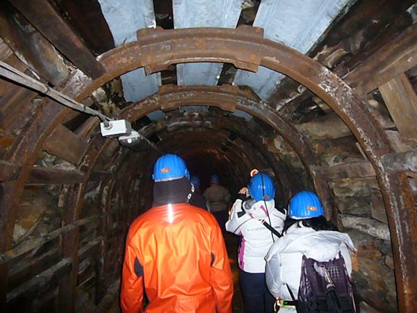 Visitors in blue helmets walk through a mine tunnel with rusty steel arches