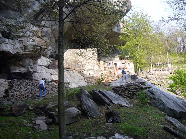 Visitors near stone walls beneath a huge overhanging rock among trees