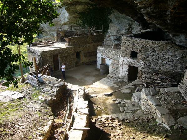 Ancient dry-stone shelters under a rock overhang, a visitor looks around
