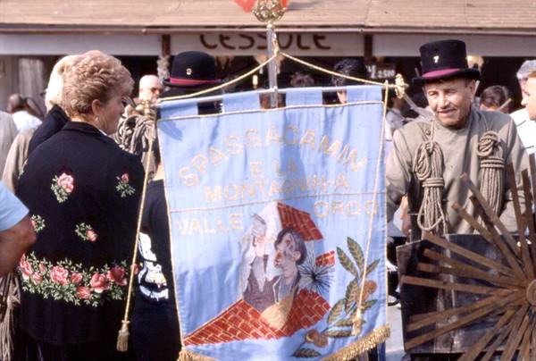 Man in black hat with ropes beside embroidered blue banner at festival