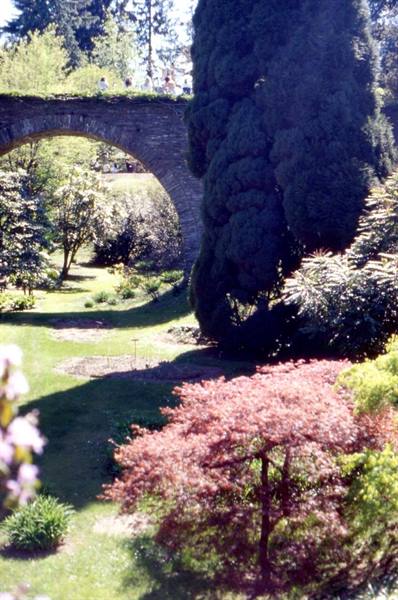 Visitors cross a stone arch bridge over sunny gardens, green and red trees below