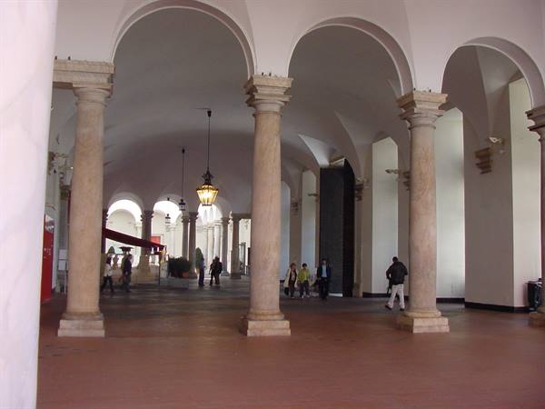 Stone columned arcade with yellow lanterns, people walking beneath vaulted ceilings