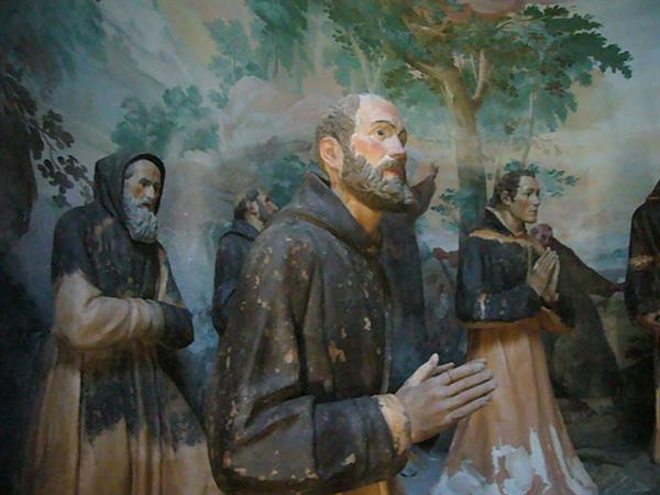 Statues of praying friars in worn brown robes before a painted tree mural
