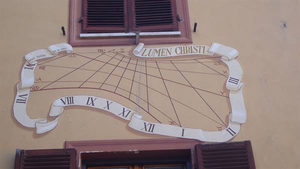 Painted sundial reading Lumen Christi between two shuttered windows
