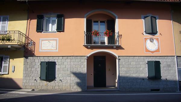 Pink facade with painted sundial, green shutters and geraniums on balcony