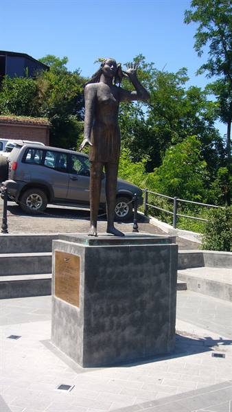 Bronze statue of a woman with raised hand on concrete pedestal beside a parked SUV