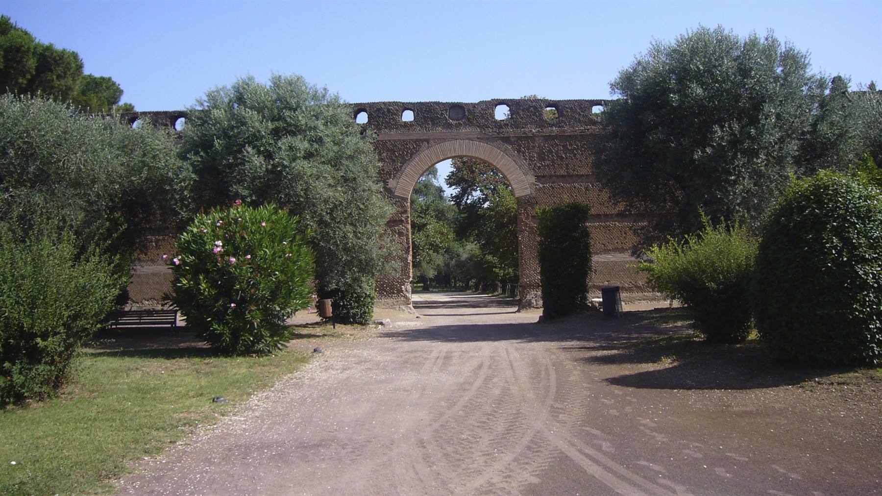 Arco in mattoni di antica cinta muraria, vialetto sterrato tra oleandri e pini, sole