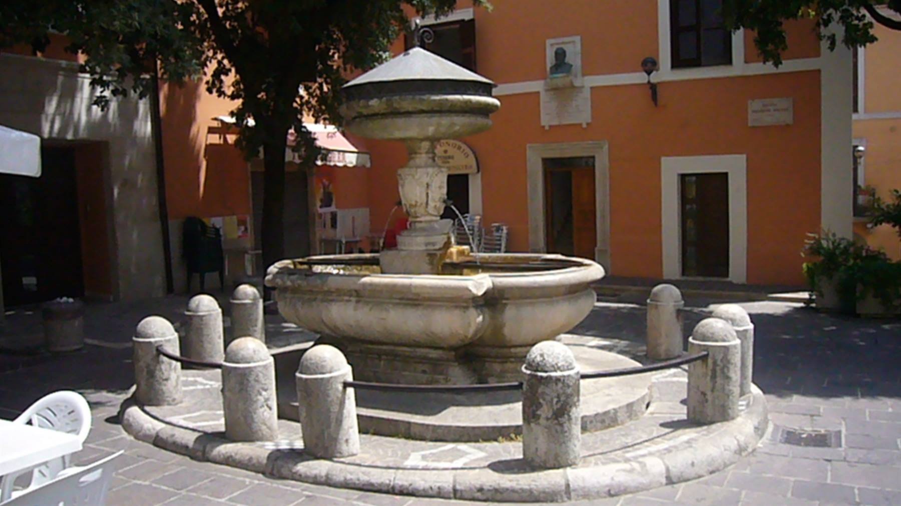 Fontana in pietra circondata da colonnine in una piazza con edifici ocra