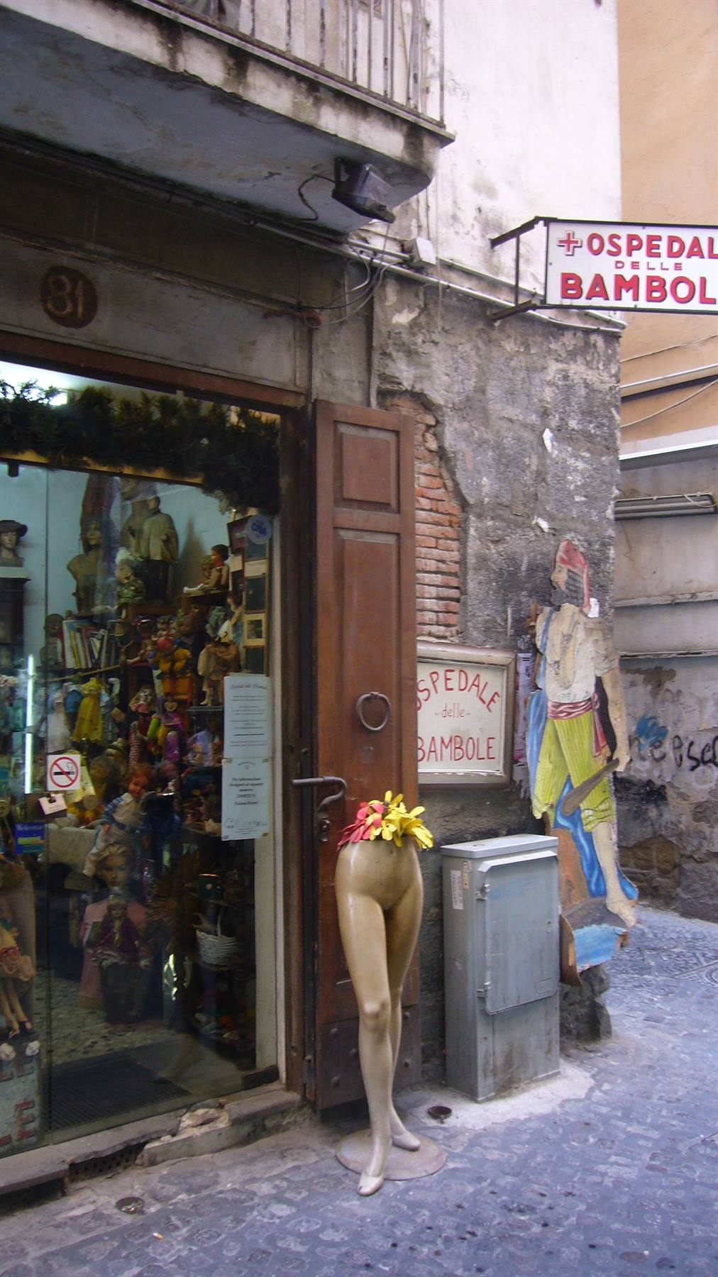 Manichino senza busto con fiori davanti a un negozio di bambole, insegna Ospedale delle Bambole
