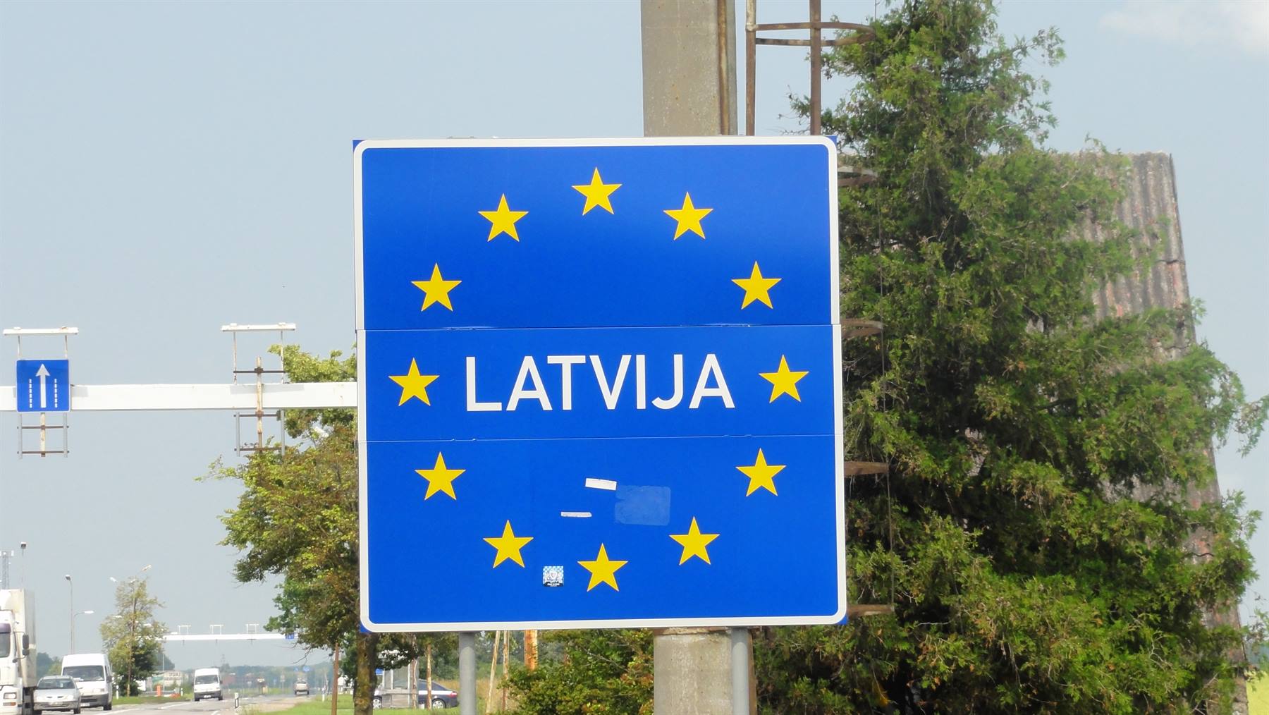 Cartello blu con stelle gialle e scritta LATVIJA al confine, auto in strada