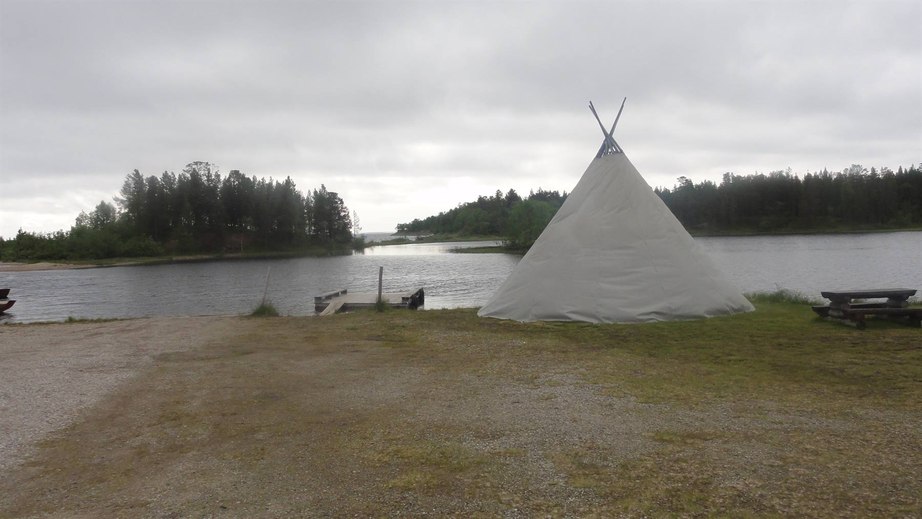 Tenda bianca a cono sulla riva erbosa di un lago con tavolo di legno