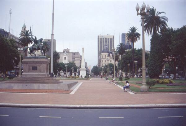 Equestrian bronze statue in a plaza, palm trees, people on benches, grey sky